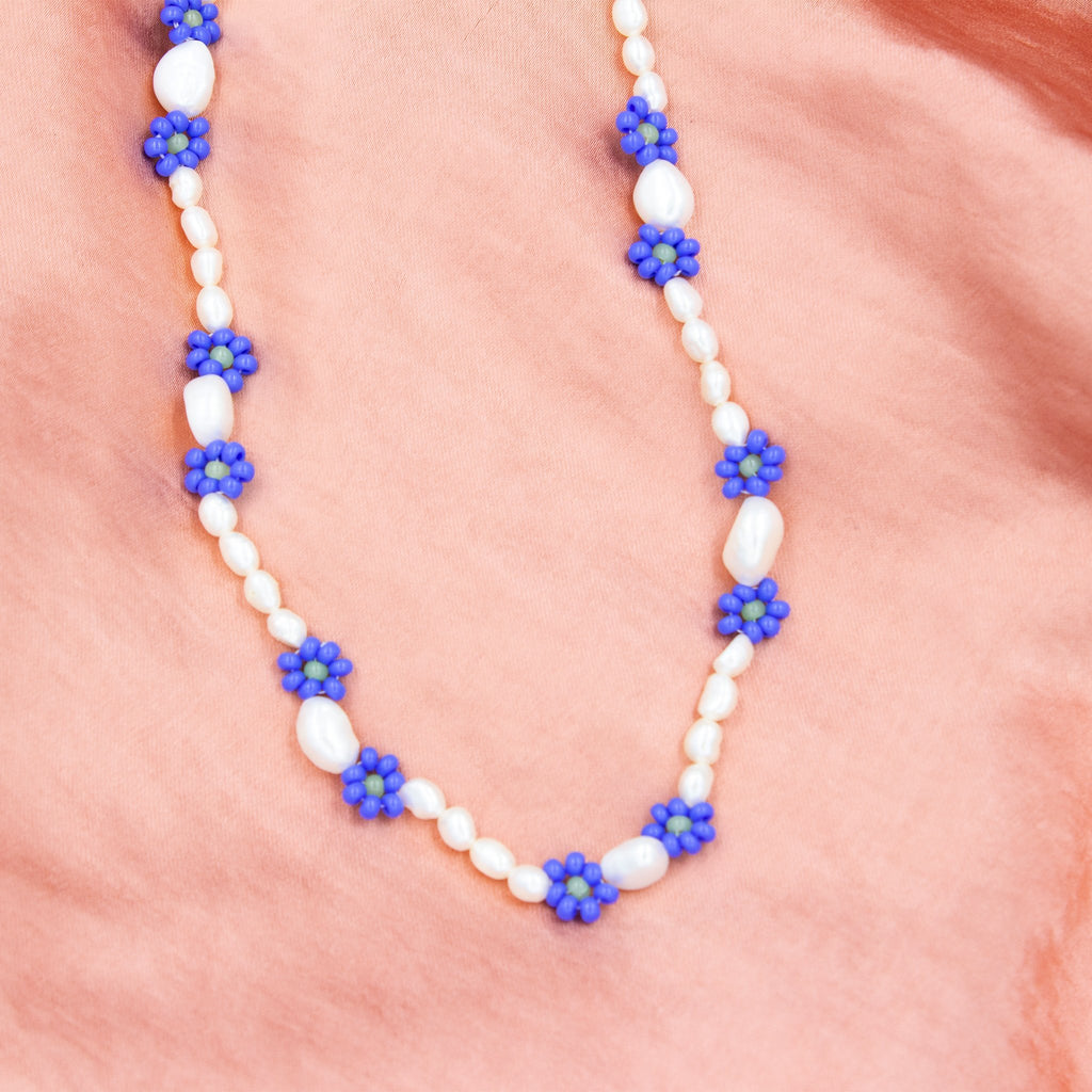 Eine handgefertigte Perlenkette mit Gänseblümchen-Motiven, gemeinsam mit einer türkischen Kunsthandwerkerin hergestellt. Die Perlen sind naturbelassen und variieren in Form, Größe und Farbe. Die Kette wird in einem kleinen Stoffbeutel zur Aufbewahrung geliefert. Der Ton ist elegant und herzlich und betont Handwerkskunst sowie kulturelle Authentizität.